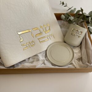 גביע קידוש עם אותיות זהב בולטות לצד כיסוי חלה לבן עם כיתוב שבת ויום טוב מוזהב